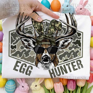May include: White t-shirt with a deer wearing a camouflage cap with bunny ears and sunglasses. The shirt displays the text "EGG HUNTER". The image is surrounded by Easter decorations, including eggs and flowers.