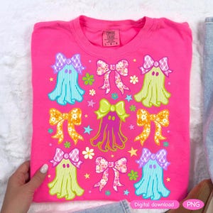 May include: A hot pink Comfort Colors t-shirt featuring a repeating pattern of cartoon ghosts with bows. The ghosts are in various colors, including blue, green, and purple, with floral and plaid bows. The design includes stars and flowers.