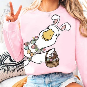 May include: Pink sweatshirt featuring a cartoon duck dressed as the Easter Bunny. The duck wears bunny ears and a floral garland with Easter eggs. It holds a basket with eggs and a chocolate bunny. The design is whimsical and festive.