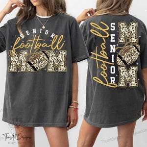 Puede incluir: Camiseta gris oscuro con "Senior Football Mom" en letras doradas y blancas. El balón de fútbol y la palabra "MOM" tienen un diseño de estampado de leopardo. La parte trasera de la camiseta repite el diseño.