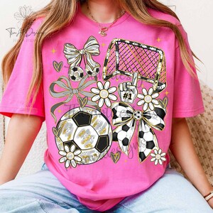May include: Pink t-shirt with a soccer-themed graphic. The design includes soccer balls, bows, a goal, daisies, and a hand with the number one. The graphic has gold and silver accents.