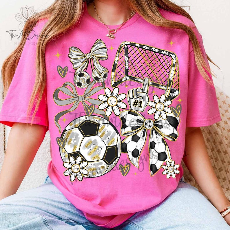 May include: Pink t-shirt with a soccer-themed graphic. The design includes soccer balls, bows, a goal, daisies, and a hand with the number one. The graphic has gold and silver accents.