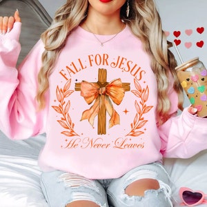 Puede incluir: Sudadera rosa con un gráfico de una cruz de madera con un lazo naranja y el texto "Fall for Jesus He Never Leaves".