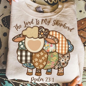 May include: A beige sweatshirt featuring a colorful patchwork sheep design. The sheep is composed of various floral and patterned fabrics. The text "The Lord Is My Shepherd" and "Psalm 23:1" are printed on the shirt.