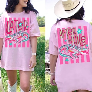 Può includere: T-shirt rosa con grafica di un alligatore e la scritta "LATER" sul davanti. Il retro della maglietta presenta le parole "AFTER WHILE" e un'altra grafica di alligatore. Il design include strisce rosa e stelle turchesi.