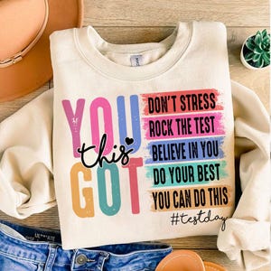 May include: A beige sweatshirt with a colorful graphic that says "You got this" and includes the text "Don't stress, Rock the test, Believe in you, Do your best, You can do this, #testday".
