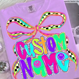 May include: A lilac Comfort Colors t-shirt featuring a large, colorful bow design with a black and white checkered pattern. The words "CUSTOM NAME" are displayed in a vibrant, bubble-style font with smiley face accents.
