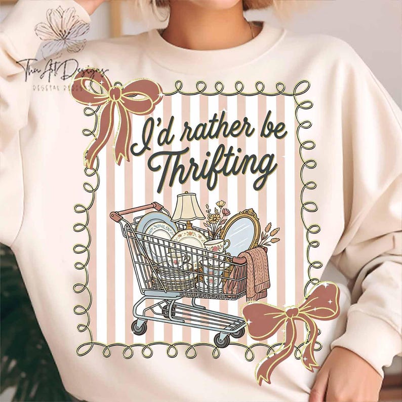 May include: Cream-colored sweatshirt with a graphic featuring a shopping cart filled with vintage items. The text "I'd rather be Thrifting" is displayed above the cart. The design includes decorative bows and a striped background.