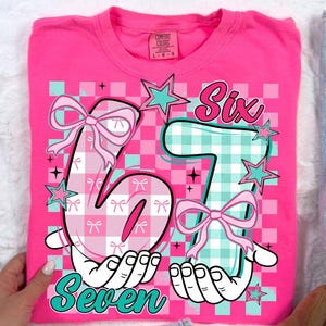 Könnte beinhalten: Ein leuchtend pinkfarbenes Comfort Colors T-Shirt mit einem grafischen Design. Das Design zeigt die Zahlen "6" und "7" in einem karierten Muster, verziert mit Schleifen und Sternen. Das Wort "Seven" steht darunter.