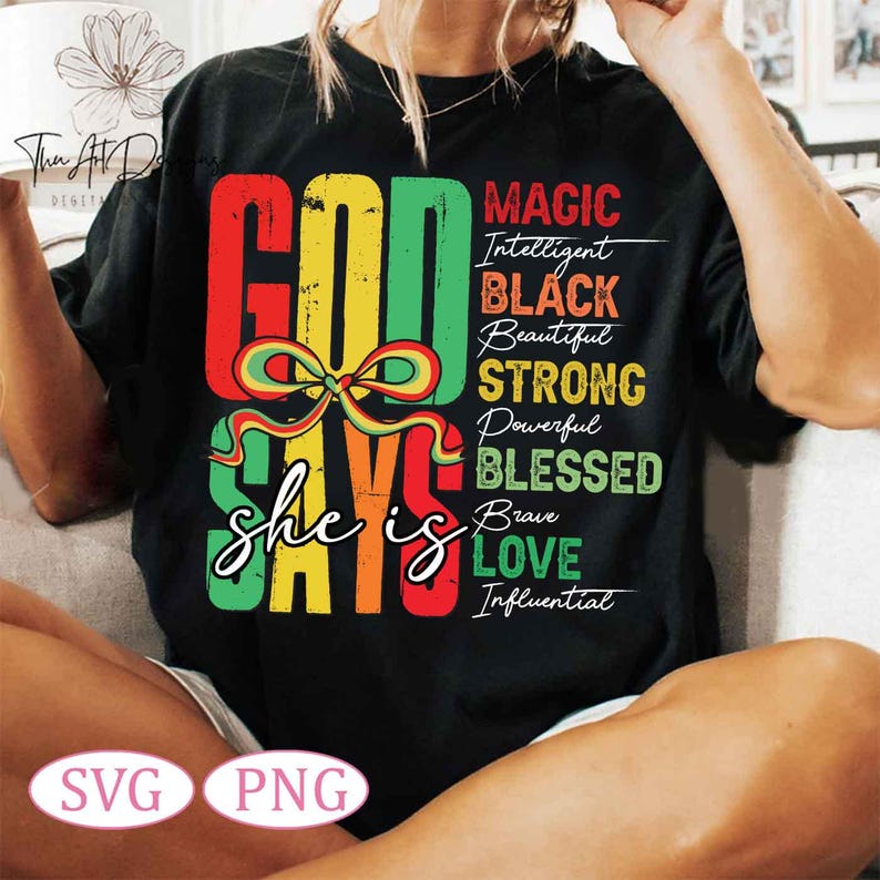 Op de afbeelding: Een zwart T-shirt met een kleurrijk grafisch ontwerp dat "God says she is" zegt met de woorden "Magic, Intelligent, Black, Beautiful, Strong, Powerful, Blessed, Brave, Love, Influential" in een verticale kolom rechts van de hoofdtekst.