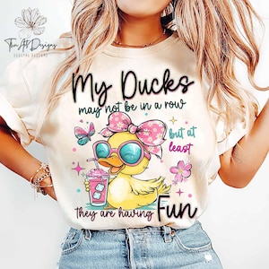 May include: Cream-colored t-shirt with a graphic of a yellow duck wearing sunglasses and a pink bow. The text reads "My Ducks may not be in a row but at least they are having Fun."