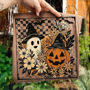 Peut inclure: Un coussin carré sur le thème d'Halloween avec un fantôme et une citrouille-lanterne, tous deux portant des chapeaux de sorcière. Le motif comprend des fleurs, des chauves-souris et un fond à carreaux dans les tons de marron, d'orange et de crème.