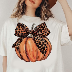 May include: White t-shirt featuring a graphic of an orange pumpkin with a brown and black leopard print bow.