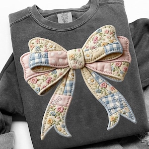 May include: A charcoal gray sweatshirt featuring a large, decorative bow. The bow is made of patchwork fabric with floral, polka dot, and gingham patterns in pastel pink, blue, and cream colors. The brand tag "Comfort Colors" is visible.