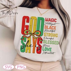 Op de afbeelding: Een wit sweatshirt met een kleurrijk grafisch ontwerp dat "God Says She Is" zegt met de woorden "Magic, Intelligent, Black, Beautiful, Strong, Powerful, Blessed, Brave, Love, Influential" in zwarte tekst.