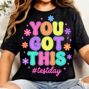 Pode incluir: Camiseta preta com a frase "YOU GOT THIS" em letras coloridas, rodeada por flores e estrelas. A hashtag "#testday" est&aacute; abaixo. Um design alegre e motivador, ideal para exames.