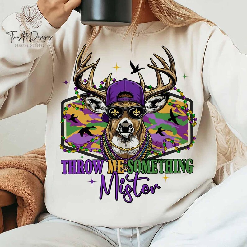 May include: Cream-colored sweatshirt featuring a deer wearing sunglasses, a purple cap, and a beaded necklace. The deer is set against a camouflage background with the text "THROW ME SOMETHING Mister".