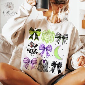 May include: Cream-colored sweatshirt featuring a collection of graphic designs. The designs include various bows in green, black, and purple, a black and white striped creature, and the text "BEETLEJUICE" in green. A crescent moon and a striped suit are also featured.