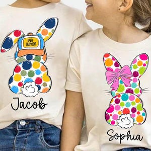Può includere: Due magliette bianche con un design di coniglio a pois colorati. Una maglietta dice "Egg Hunter" sul cappello del coniglio e "Jacob" sotto il coniglio. L'altra maglietta ha un fiocco rosa sul coniglio e dice "Sophia" sotto il coniglio.