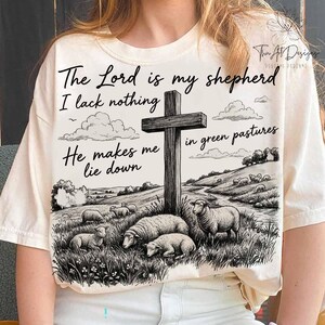 May include: Cream-colored t-shirt featuring a black and white pastoral scene with sheep, a cross, and the text "The Lord is my shepherd, I lack nothing, He makes me lie down in green pastures." The design includes clouds and a landscape.