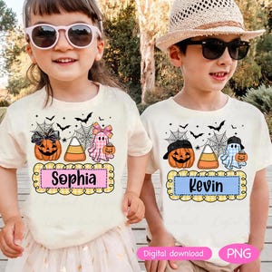以下が含まれることがあります： ハロウィーンをテーマにしたグラフィックがプリントされたクリーム色のTシャツを着た2人の子供たち。シャツにはカボチャ、お化け、コーンキャンディーが描かれており、「Sophia」と「Kevin」の名前が装飾フレームに入っています。コウモリとクモの巣がデザインを完成させています。