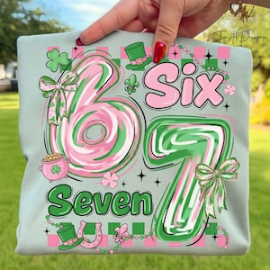 Pode incluir: Uma camiseta verde claro com os números "67" e as palavras "Six" e "Seven" em rosa e verde. O design inclui trevos, laços, um pote de ouro, uma ferradura e um chapéu de duende. A camiseta é segurada por uma pessoa.