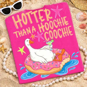 Könnte beinhalten: Ein leuchtend pinkfarbenes T-Shirt mit dem gelben Schriftzug "Hotter Than A Hoochie Coochie". Das Design zeigt eine Cartoon-Gans, die auf einem Donut-Schwimmer entspannt und einen Cocktail hält. Sonnenbrille und Muscheln sind im Hintergrund zu sehen.