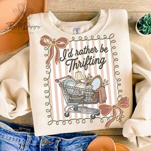 May include: Cream-colored sweatshirt with the text "I'd rather be Thrifting" and a shopping cart graphic filled with vintage items. The design includes pink and white stripes and decorative bows.