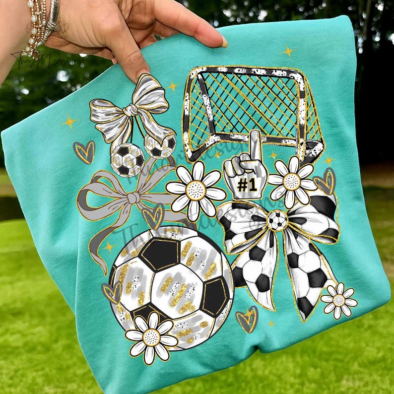 May include: Teal t-shirt featuring a soccer-themed design. The graphic includes soccer balls, bows, daisies, a soccer goal, and a hand with the number one. The design incorporates black, white, gold, and gray elements.