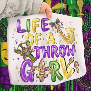 May include: White fabric with the text "LIFE OF A THROW GIRL" in purple, yellow, and green. The design includes a saxophone, crawfish, fleur-de-lis, and maracas, all in festive colors. The design is held by a person.