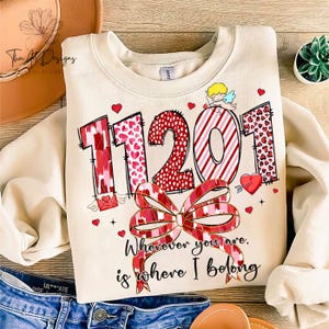Puede incluir: Una sudadera beige con un diseño gráfico rojo, rosa y blanco con los números "11201" y un lazo. El texto "Wherever you are, is where I belong" está impreso debajo de los números.