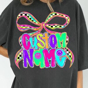 May include: Dark gray t-shirt featuring a colorful graphic design. The design includes the words "CUSTOM NAME" in vibrant, bubble letters, accented with smiley faces and a bow. The bow has a checkered pattern.