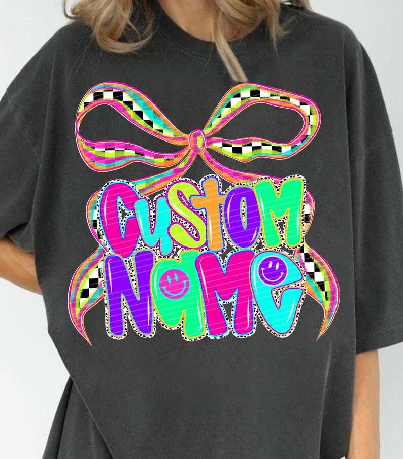 May include: Dark gray t-shirt featuring a colorful graphic design. The design includes the words "CUSTOM NAME" in vibrant, bubble letters, accented with smiley faces and a bow. The bow has a checkered pattern.
