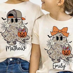 May include: Two off-white t-shirts with Halloween ghost designs. One shirt features a ghost wearing a cap and holding a pumpkin basket, with the name "Mathew" below. The other shirt has a ghost with a bow and the name "Emily".