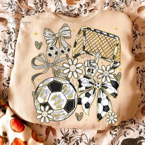 May include: Beige sweatshirt with a soccer theme. The design includes soccer balls, bows, a goal, daisies, and a hand with the number one. The graphics are in black, white, and gold.