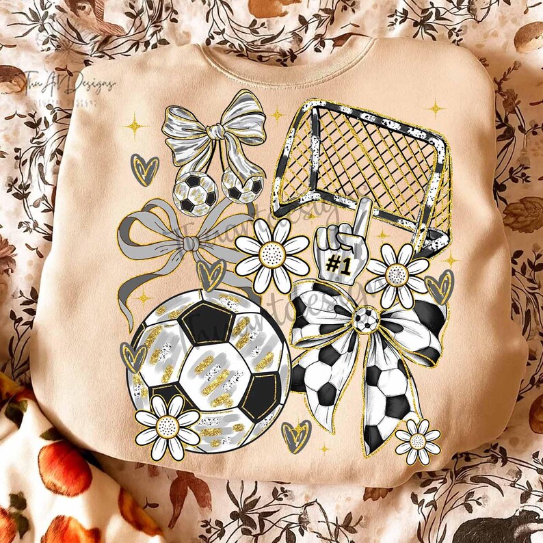 May include: Beige sweatshirt with a soccer theme. The design includes soccer balls, bows, a goal, daisies, and a hand with the number one. The graphics are in black, white, and gold.
