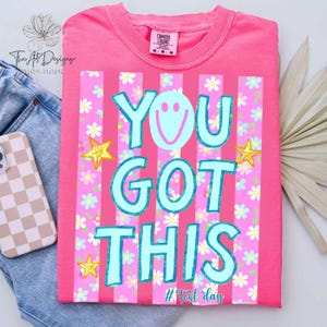 May include: A pink t-shirt with the text "YOU GOT THIS" in light blue, featuring a smiley face and star graphics. The shirt has vertical pink stripes and a floral pattern. The tag reads "Comfort Colors".