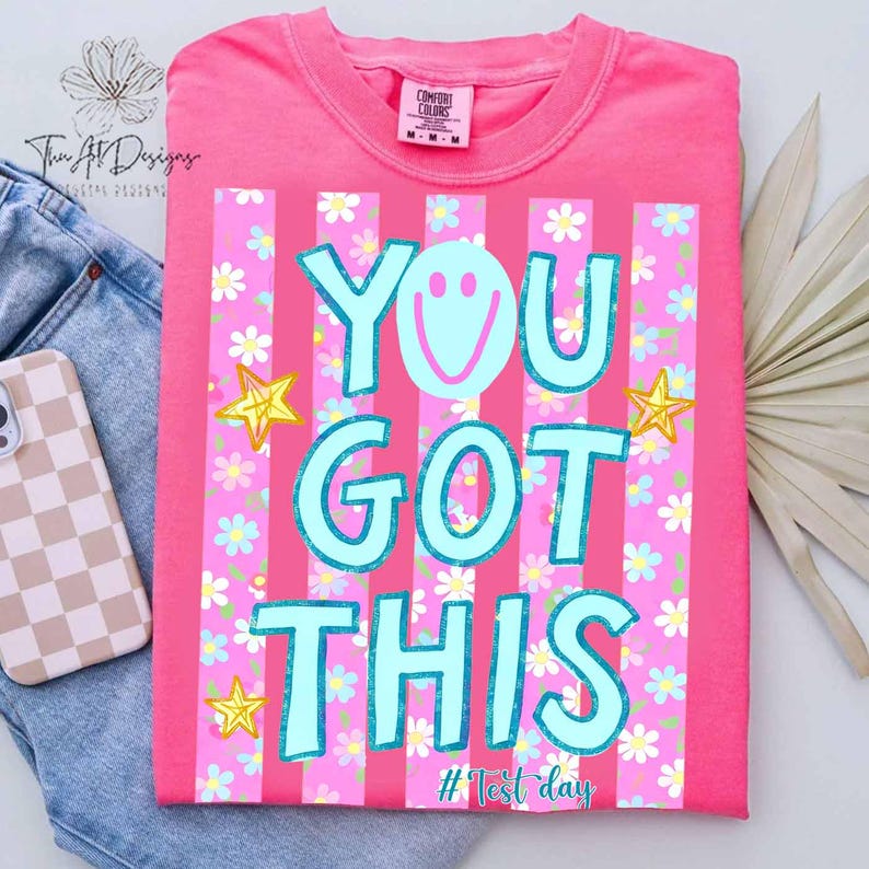 May include: A pink t-shirt with the text "YOU GOT THIS" in light blue, featuring a smiley face and star graphics. The shirt has vertical pink stripes and a floral pattern. The tag reads "Comfort Colors".