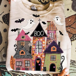 Puede incluir: Sudadera beige con un diseño de casas de Halloween coloridas y la palabra "BOO". El diseño incluye casas con varios patrones, murciélagos y fantasmas. La sudadera está doblada y sostenida por manos.