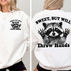 May include: White sweatshirt with a raccoon graphic and the text "Sweet, But Will Throw Hands." The raccoon is depicted with its hands up. The design is on the front and back of the sweatshirt.