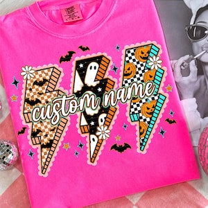 May include: A vibrant pink t-shirt with three lightning bolt designs, each decorated with Halloween motifs. The bolts feature bats, ghosts, pumpkins, and the text "custom name". The shirt has a Comfort Colors label.