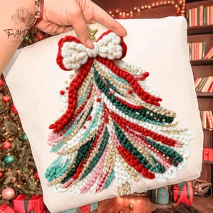 Op de afbeelding: Een vierkant, crèmekleurig decoratief kussen met een kerstboomontwerp. De boom is gemaakt van verschillende kleurrijke garens, waaronder rood, groen en wit, met gouden accenten en een strik aan de bovenkant. Het kussen wordt door een hand vastgehouden.
