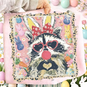 May include: A pink blanket featuring a raccoon wearing heart-shaped sunglasses and a bow, with bunny ears. The design includes Easter eggs, carrots, and flowers, with a leopard print border. The raccoon is blowing a bubble.