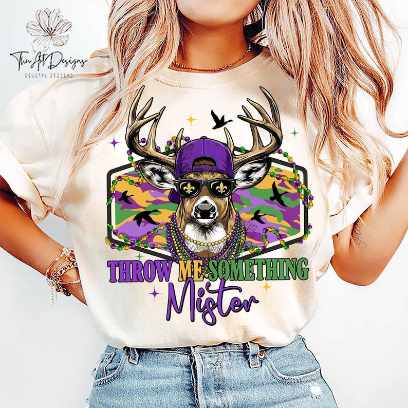 May include: Cream-colored t-shirt featuring a deer wearing a purple cap, sunglasses, and a beaded necklace. The deer is inside a green and purple camouflage design with the text "THROW ME SOMETHING Mister".