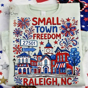 Könnte beinhalten: Hellgrünes T-Shirt mit patriotischem Design. Das Design enthält die Wörter "SMALL TOWN FREEDOM" und "RALEIGH, NC" in Rot und Blau, zusammen mit Illustrationen von Feuerwerk, Gebäuden und Sternen.