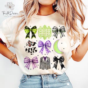 May include: Beige t-shirt featuring a collage of Halloween-themed graphics. Designs include bows in green, black, and purple, a striped snake, a pinstripe suit, a green crescent moon, and the text "BEETLEJUICE".