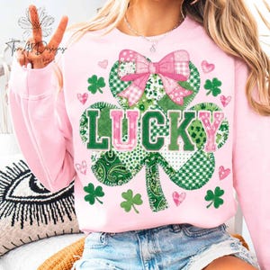 May include: A light pink sweatshirt with a large shamrock design. The shamrock is made of green and white patterned fabric, with the word "LUCKY" in green and pink. Pink hearts and green shamrocks are scattered around the design.