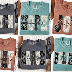 May include: Six t-shirts in different colours, each with a word in large, patterned letters. The words are "AMEN," "HOPE," "JOY," "PRAY," "GRACE," and "BELIEVE." Each shirt also has a corresponding Bible verse printed below the word.