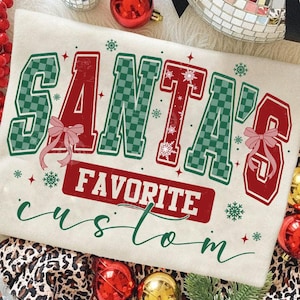 May include: Cream-colored t-shirt with the text "SANTA'S FAVORITE custom" in red and green. The letters are outlined in green and have a checkered pattern. Pink bows and snowflakes add festive details.