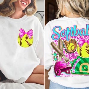 May include: White sweatshirt featuring a softball design. The front has a yellow softball with a pink bow. The back has a larger design with the word "Softball", a bat, glove, helmet, and a softball with a pink bow.